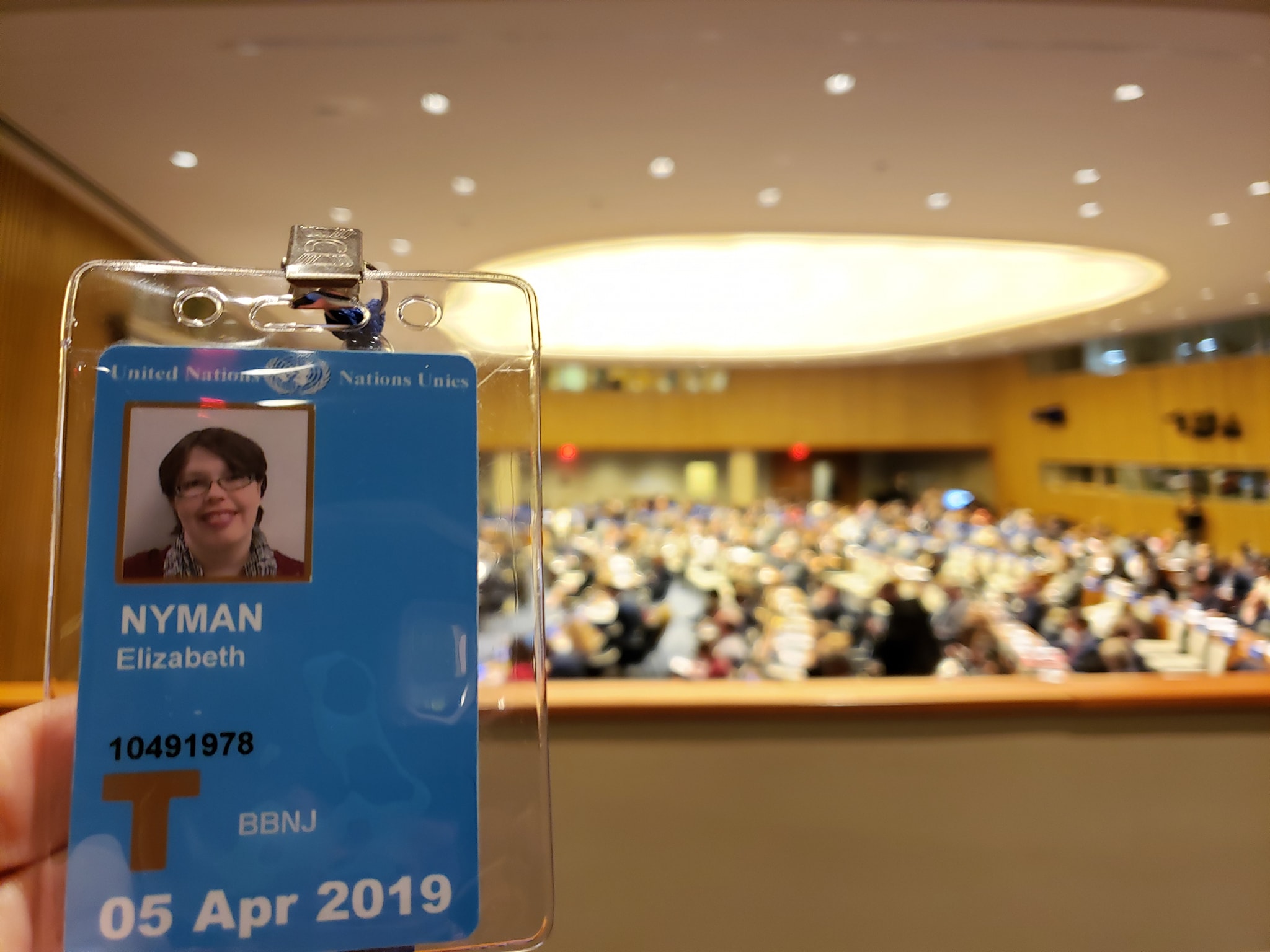 A United Nations badge for Elizabeth Nyman is held up over a background of UN negotiators.